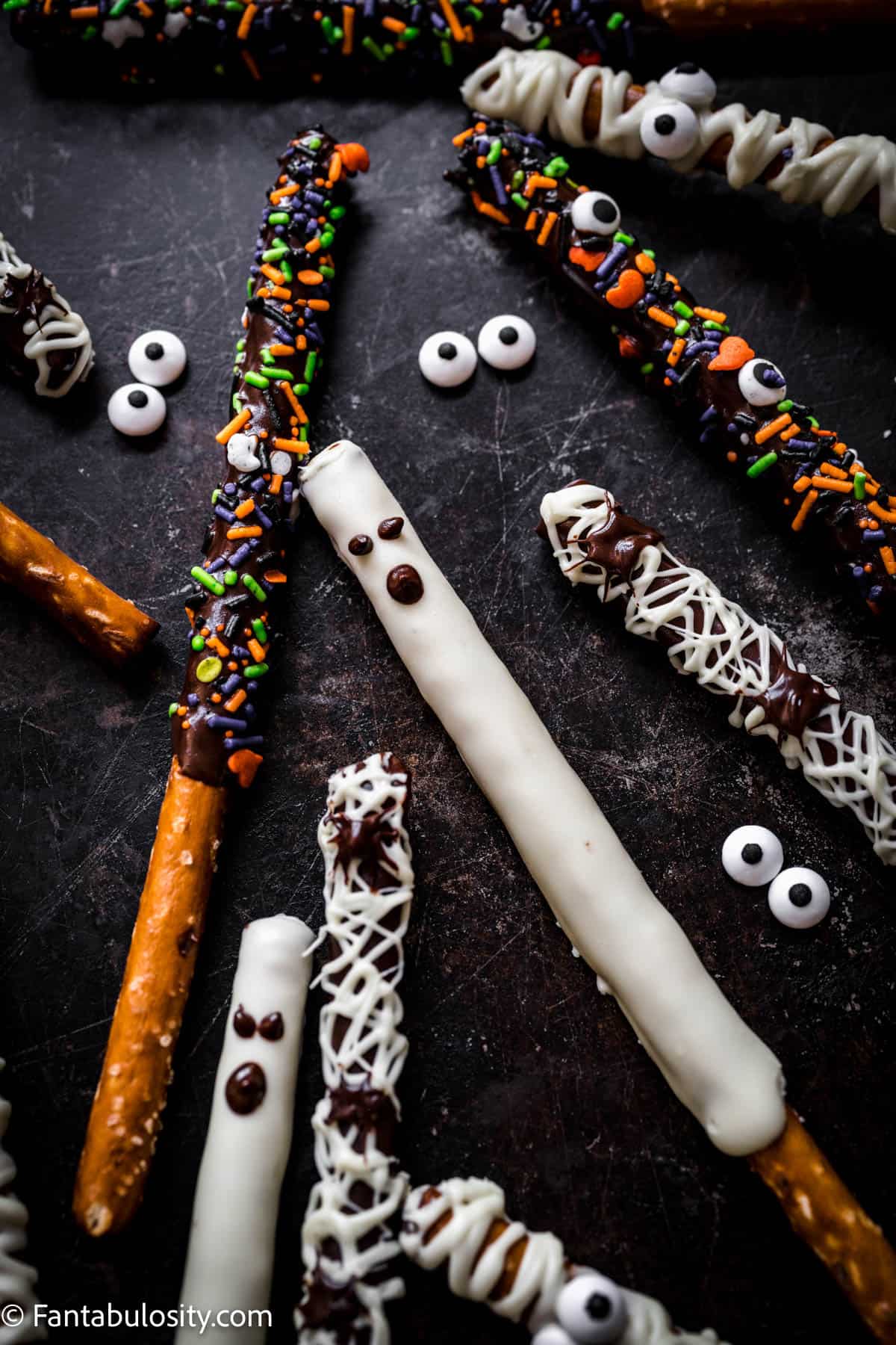Assorted chocolate dipped pretzel rods decorated in several ways for Halloween are scattered on a black background