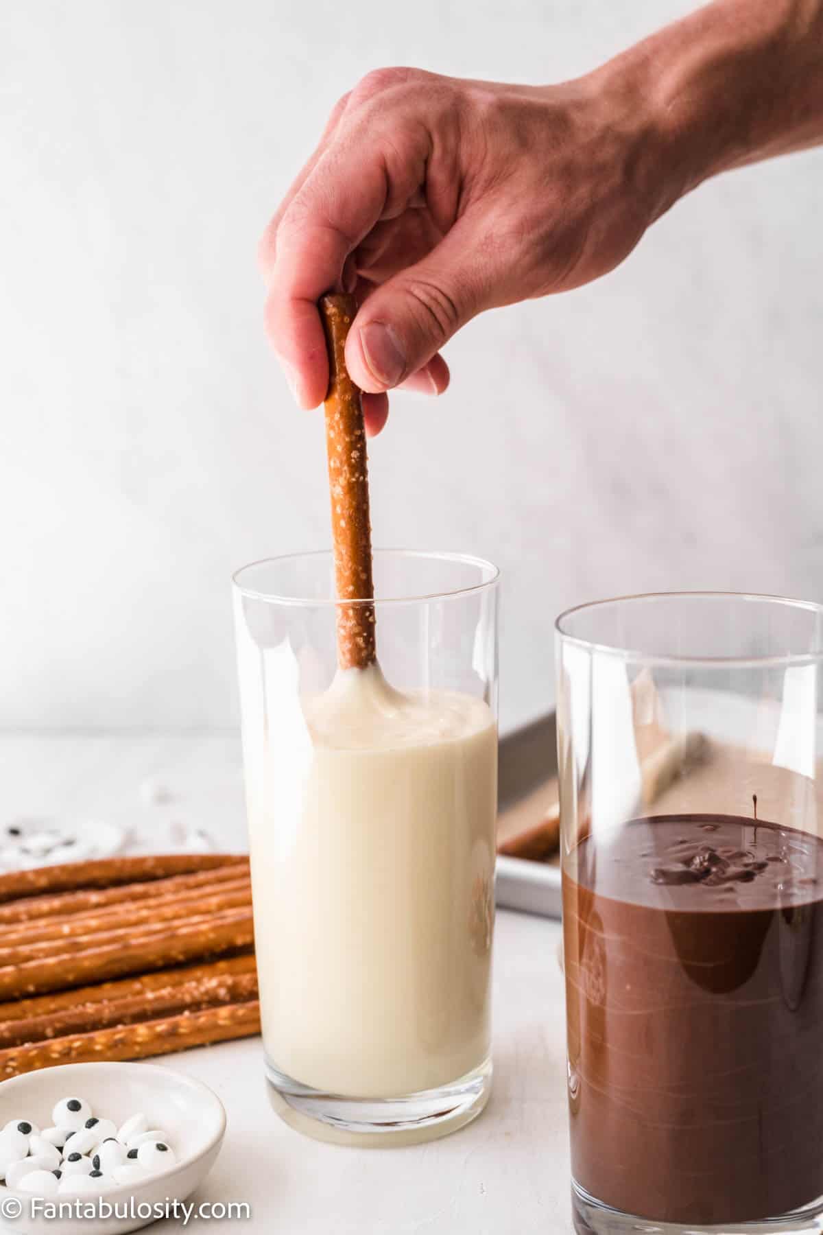 A hand holding a prezel rod is dipping it in a tall thin glass filled with melted white chocolate. A glass of melted dark chocolate, candy eyes and pretzel rods are also in the photo
