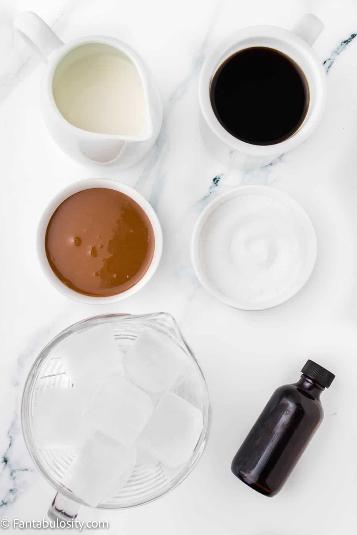 The ingredients to make Iced Caramel Lattes are displayed in various sizes of bowls on a white marble background