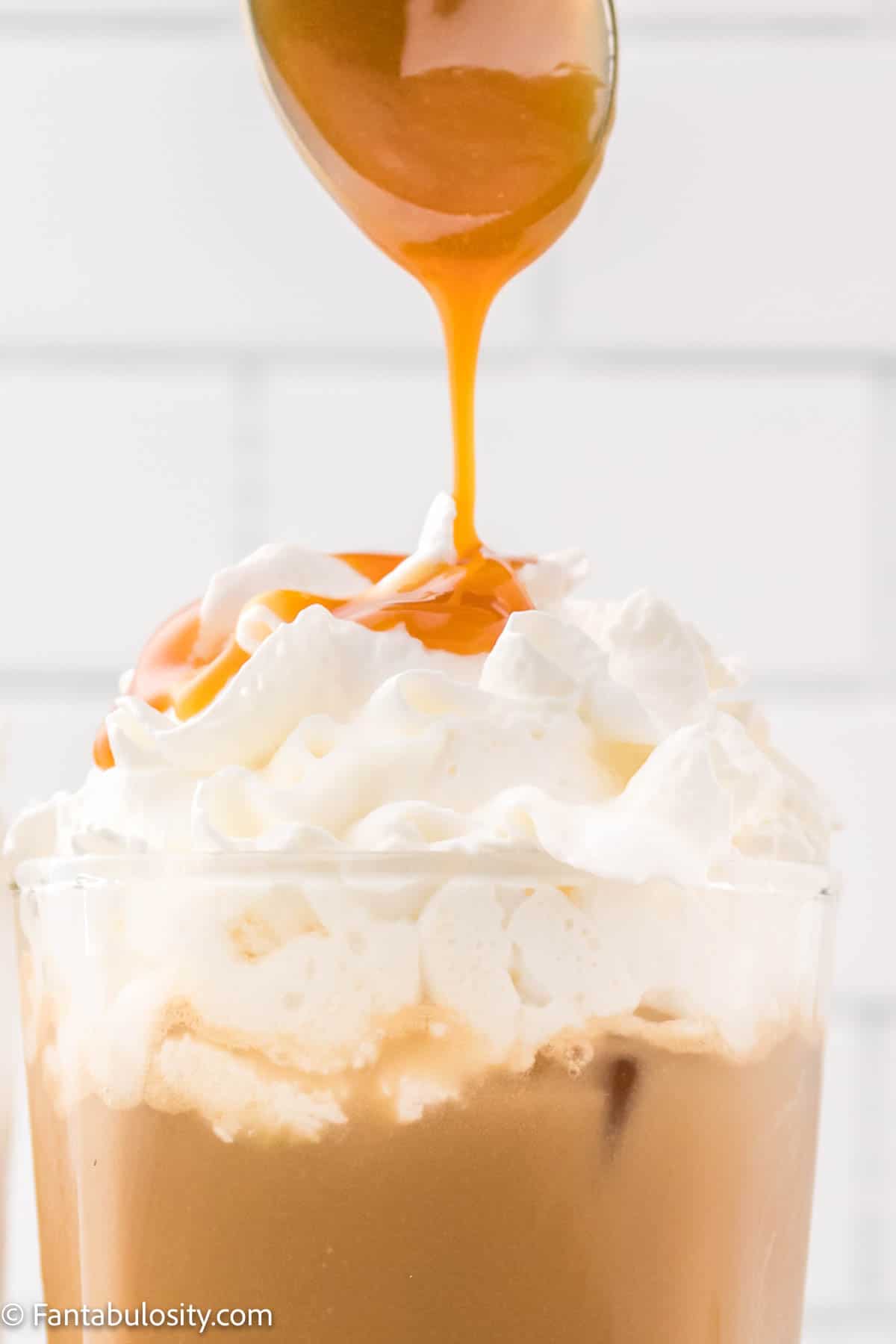 Caramel from a spoon is being drizzle over the top of a glass of iced coffee topped with whipped cream