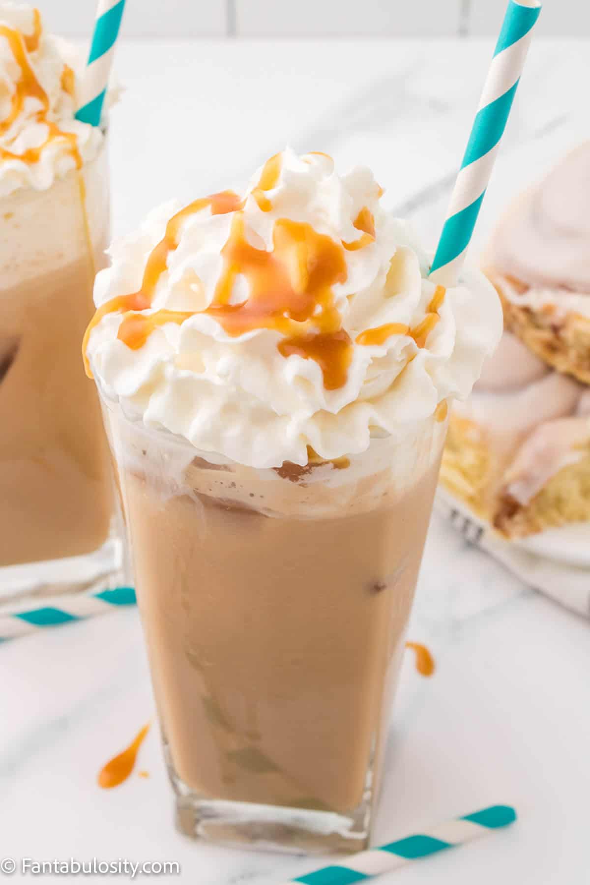 Close up photo of a glass of iced caramel latte, topped with whipped cream and caramel with a teal and white striped straw
