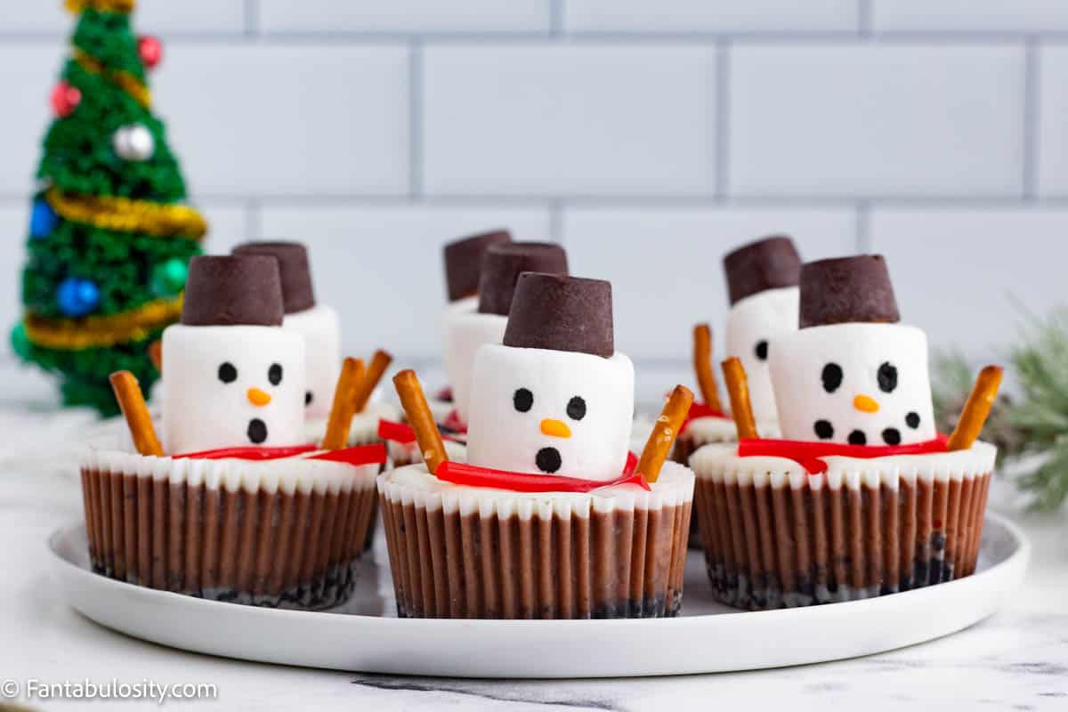 A white plates holds several chocolate cheesecake cups that are decorated to look like melting snowmen. In the background, a small Christmas tree sits off to the left