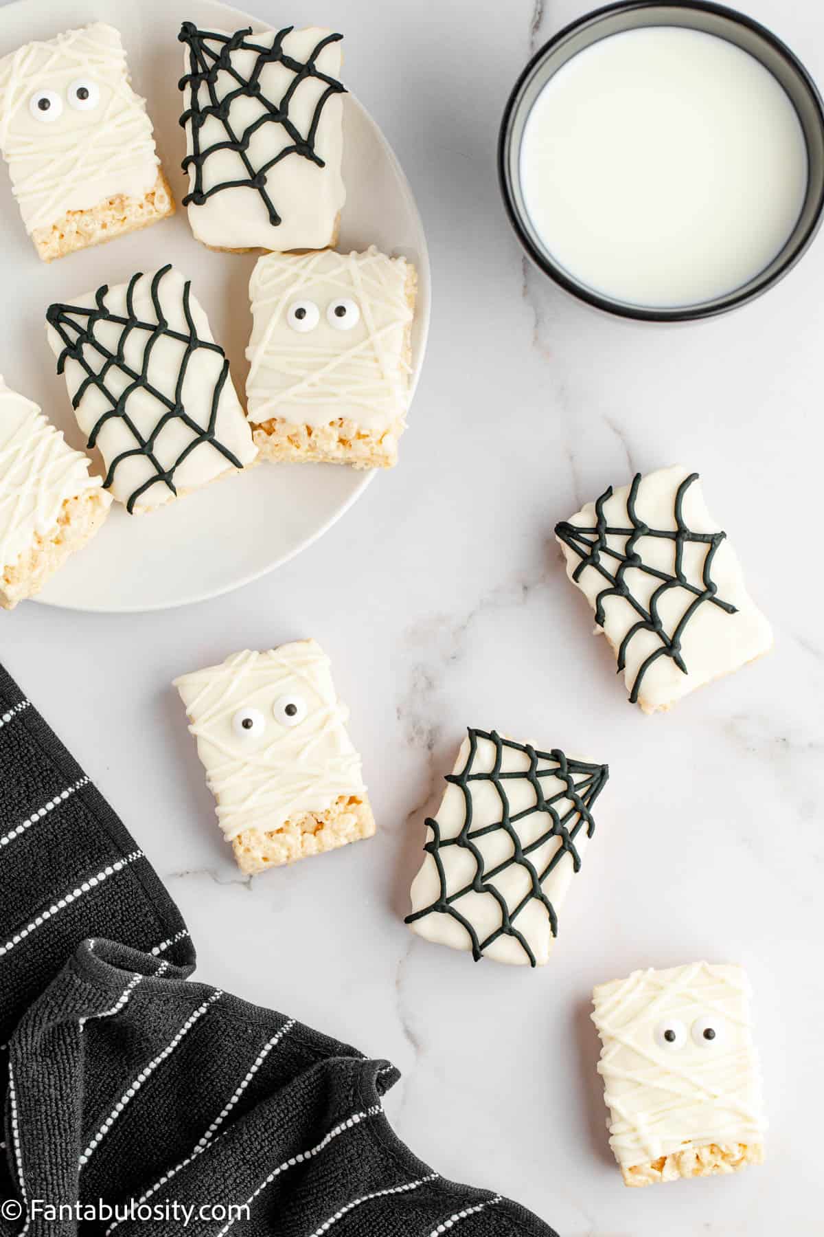 Rice krispie treats decorated to look like mummies and spider webs are displayed on a marble surface accented by a black and white towel