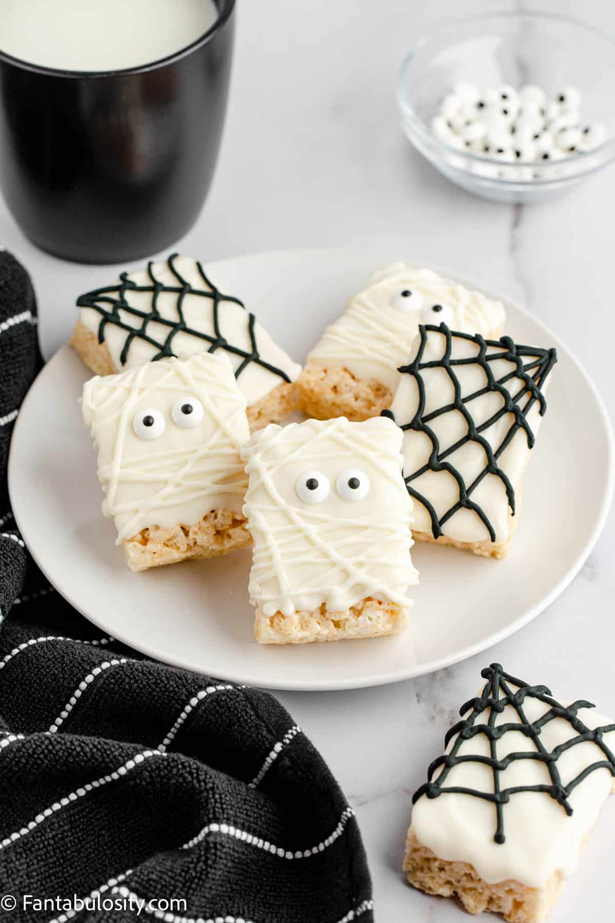 A plate full of Mummy Rice Krispie Treats is displayed with a glass of milk and another krispie treat is in the foreground beside a decorative black and white towel