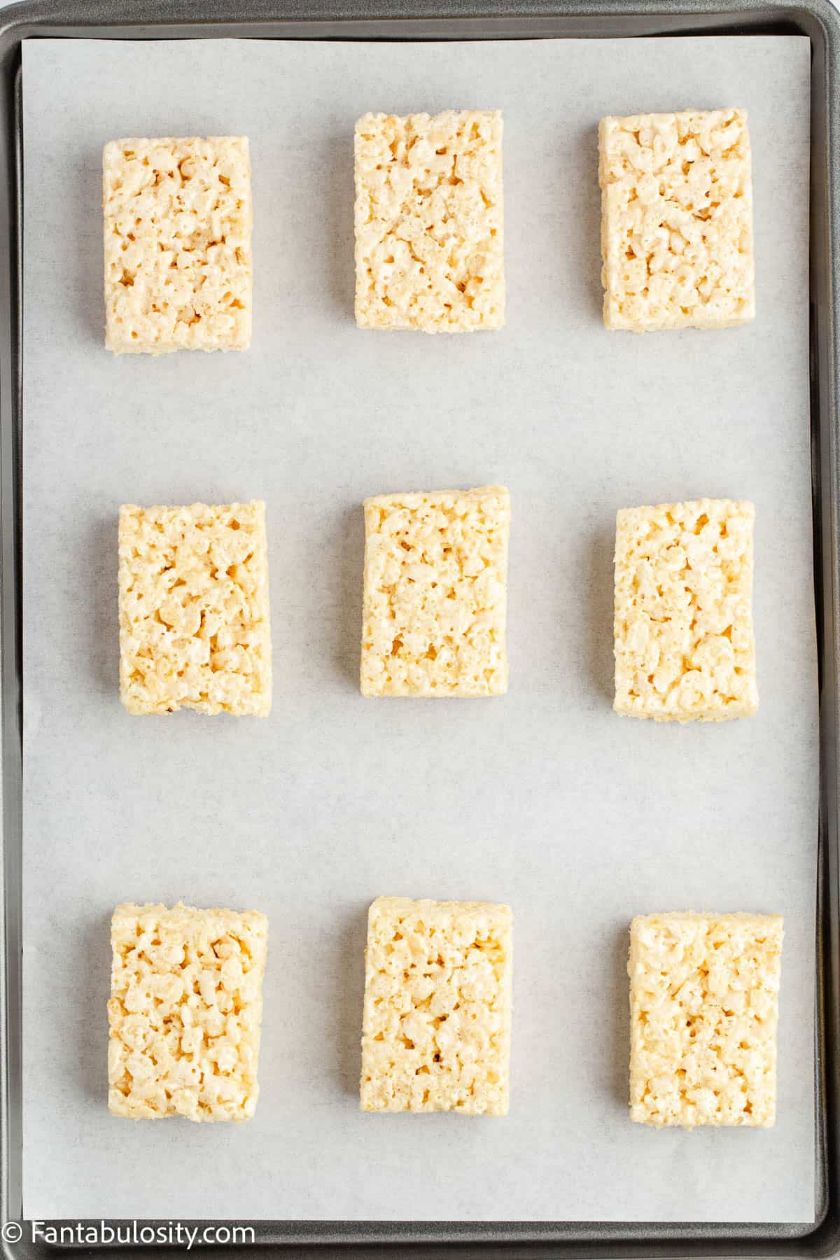 Nine unwrapped rice krispie treats are arranged on a parchment lined baking sheet