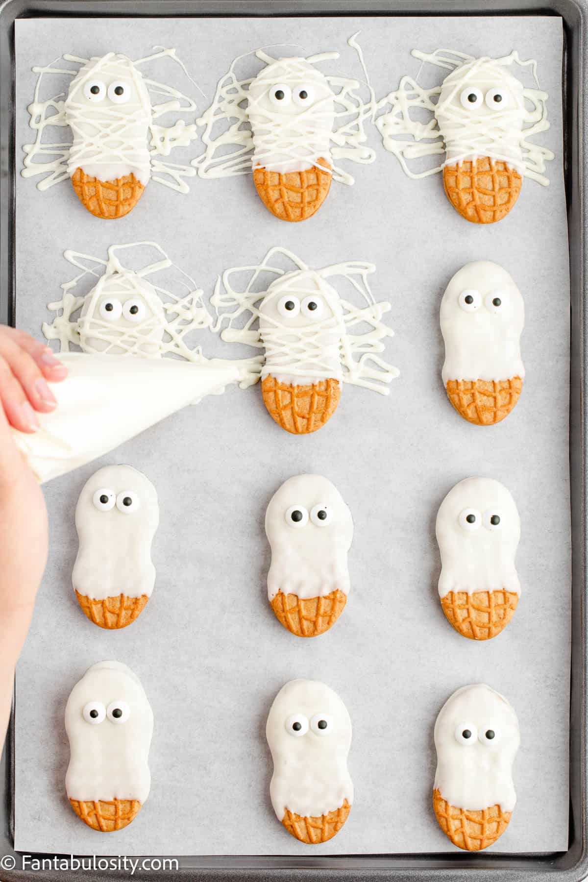 A tray of Nutter Butter Mummies cookies are displayed and a hand is drizzling melted white chocolate on the cookies for the mummy bandages