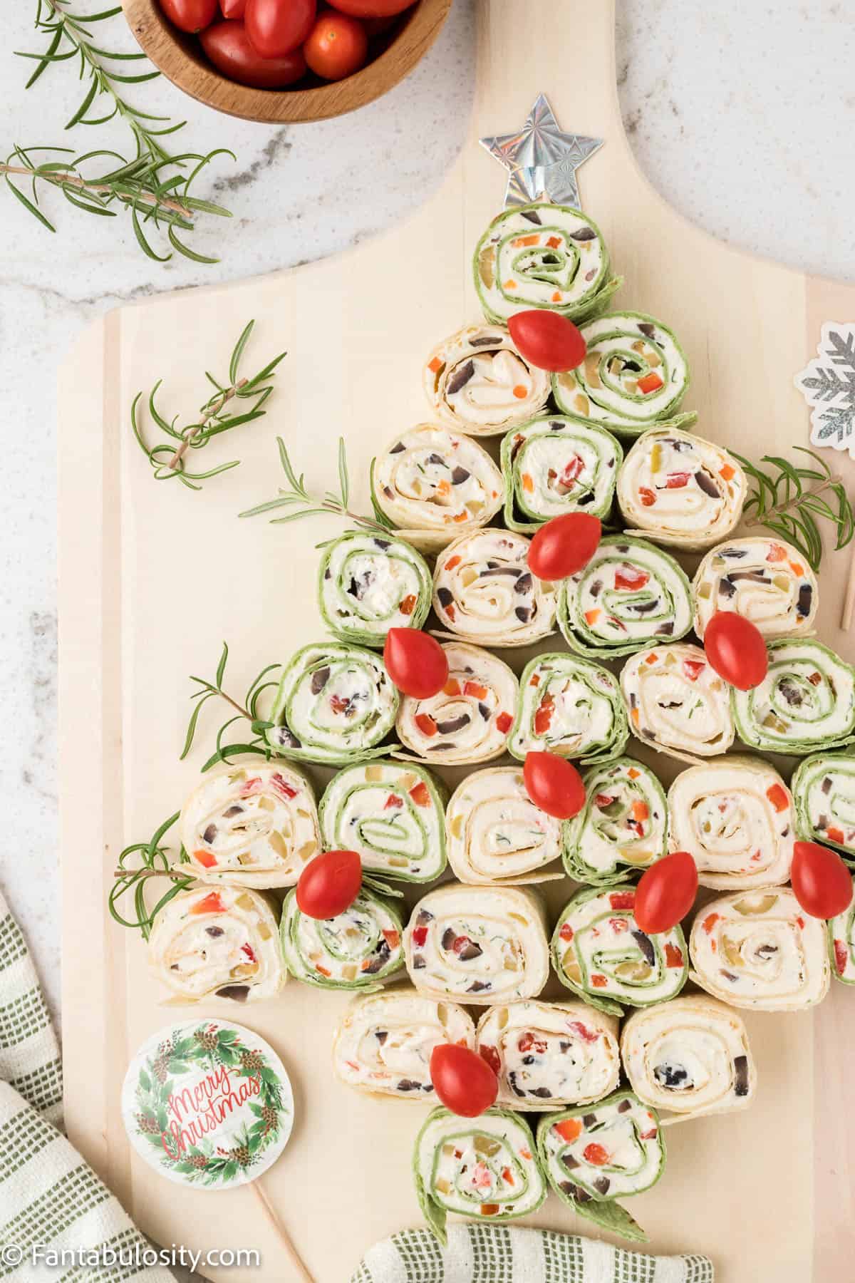 Slices of a rolled tortilla pinwheel are arranged to look like a Christmas tree, cherry tomatoes are placed to look like ornaments and there are decorative holiday picks for serving
