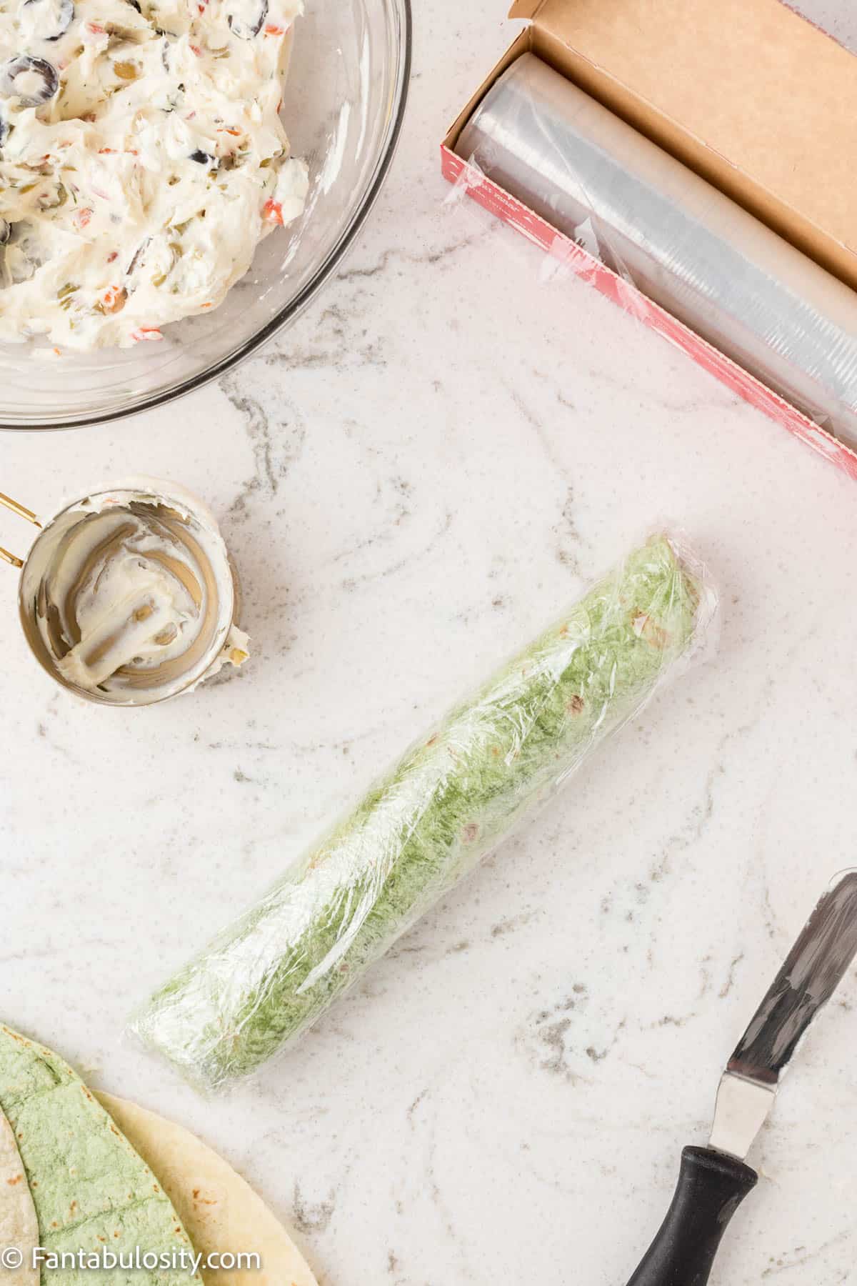 A spinach tortilla spread with a savory olive cream cheese mixture has been rolled up tightly and wrapped in plastic wrap