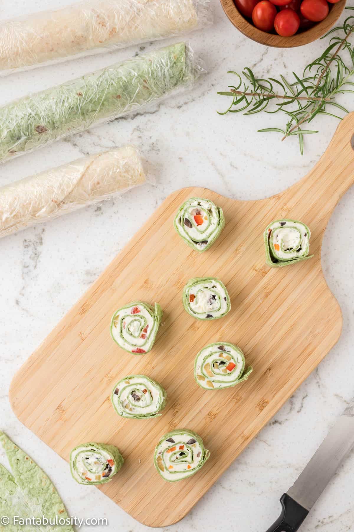 Slices of a rolled tortilla pinwheel are shown on a wooden cutting board