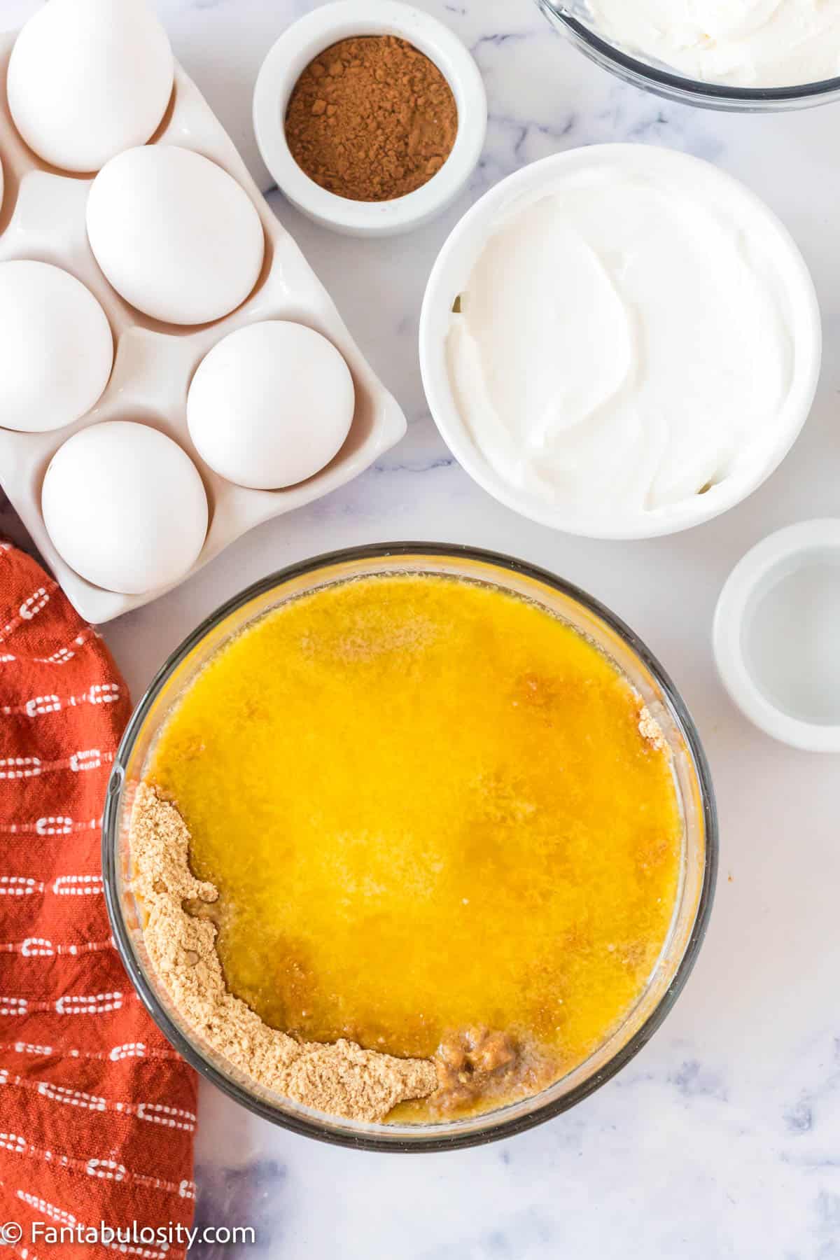 Surrounded by other baking ingredients, a large glass mixing bowl holds graham cracker crumbs and melted butter