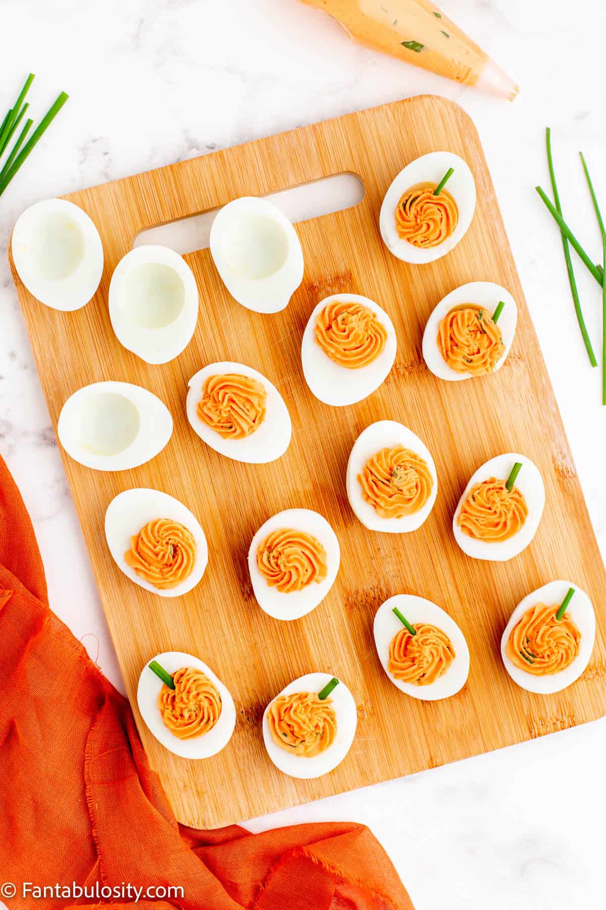 A wooden cutting board displays halved hard boiled eggs, some with piped yolk filling, are in various stages of assembly of Pumpkin Deviled Eggs