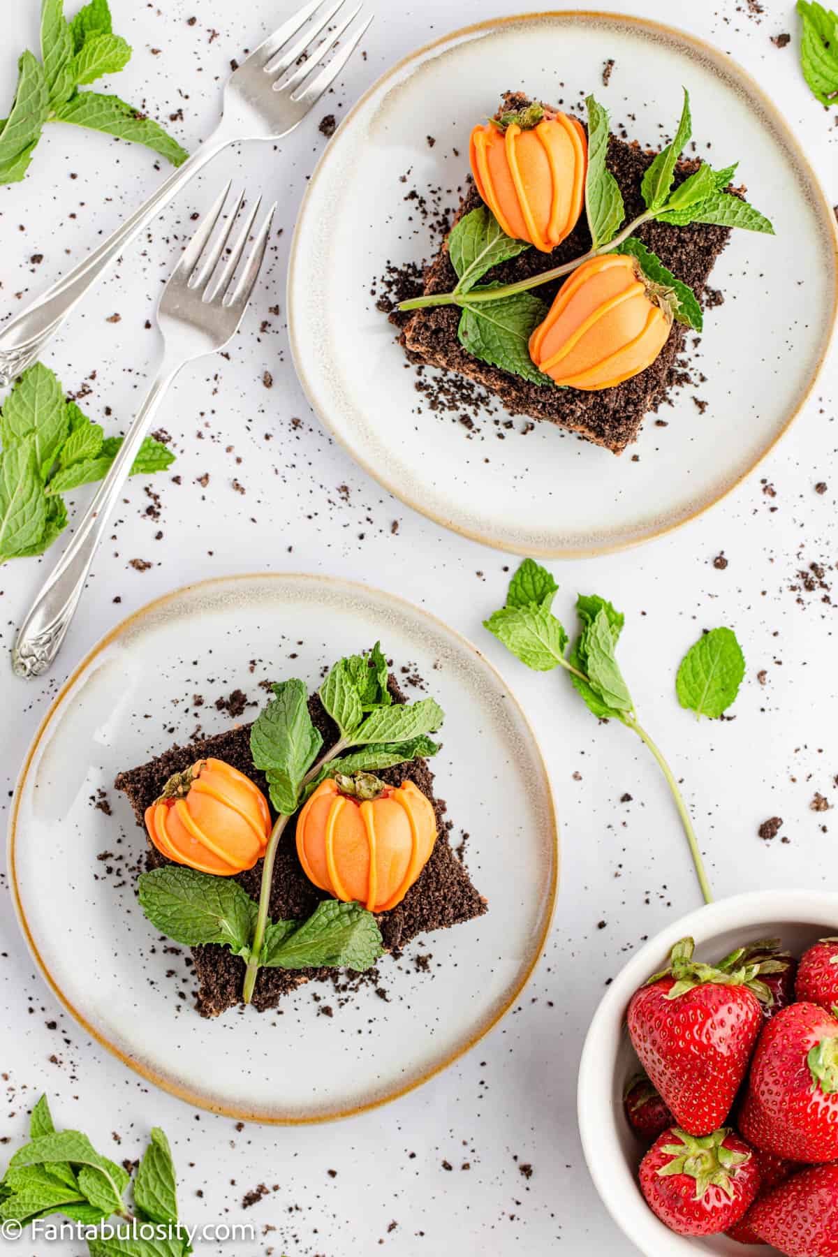 Brownies topped with chocolate covered strawberry pumpkins are plated on white plates surrounded by fresh mint and a bowl full of bright red strawberries