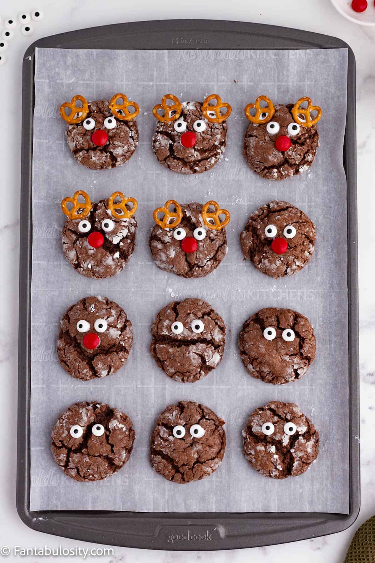 A metal baking sheet lined with parchment paper holds 12 round chocolate cookies in various stages of being decorated to look like reindeer
