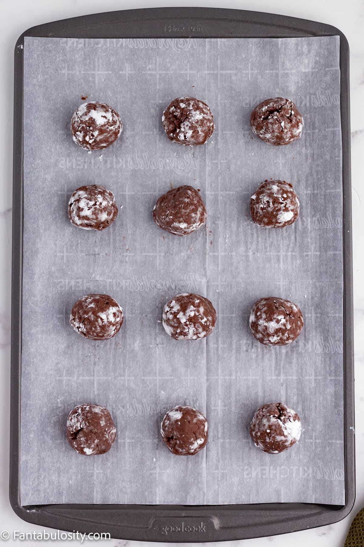 A metal baking sheet lined with parchment paper holds 12 ball of chocolate cookie dough that have been rolled in powdered sugar