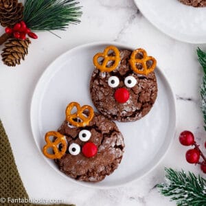 Round chocolate cookies have been decorated with candy eyes, a red candy and small pretzel twists to look like reindeer