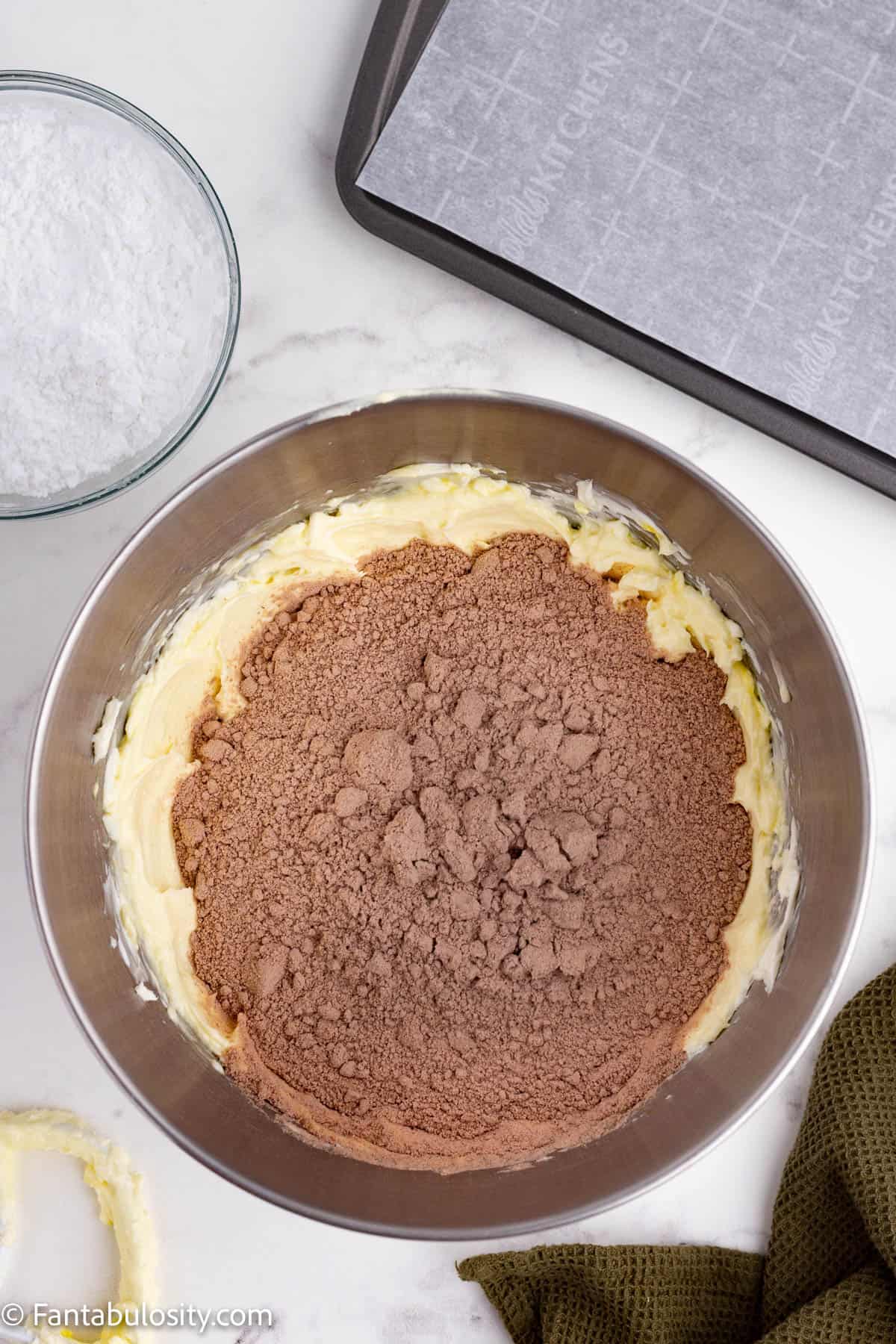 A boxed chocolate cake mix has been added to a light yellow creamy mixture in a large metal mixing bowl