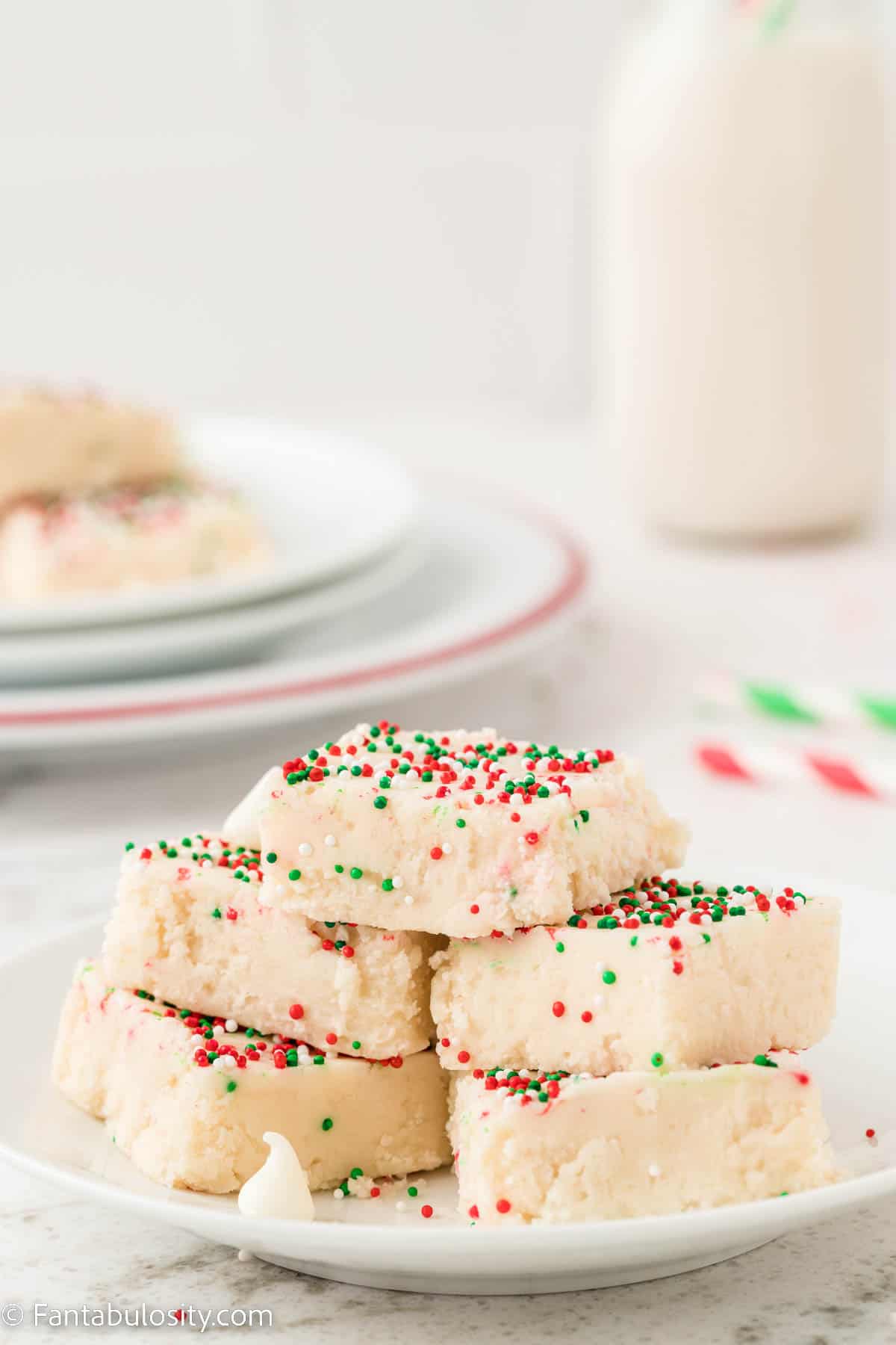 A white plate holding pieces of white fudge topped with red, white and green sprinkles is in the center of the photo and another plate of fudge and a milk bottle are in the background