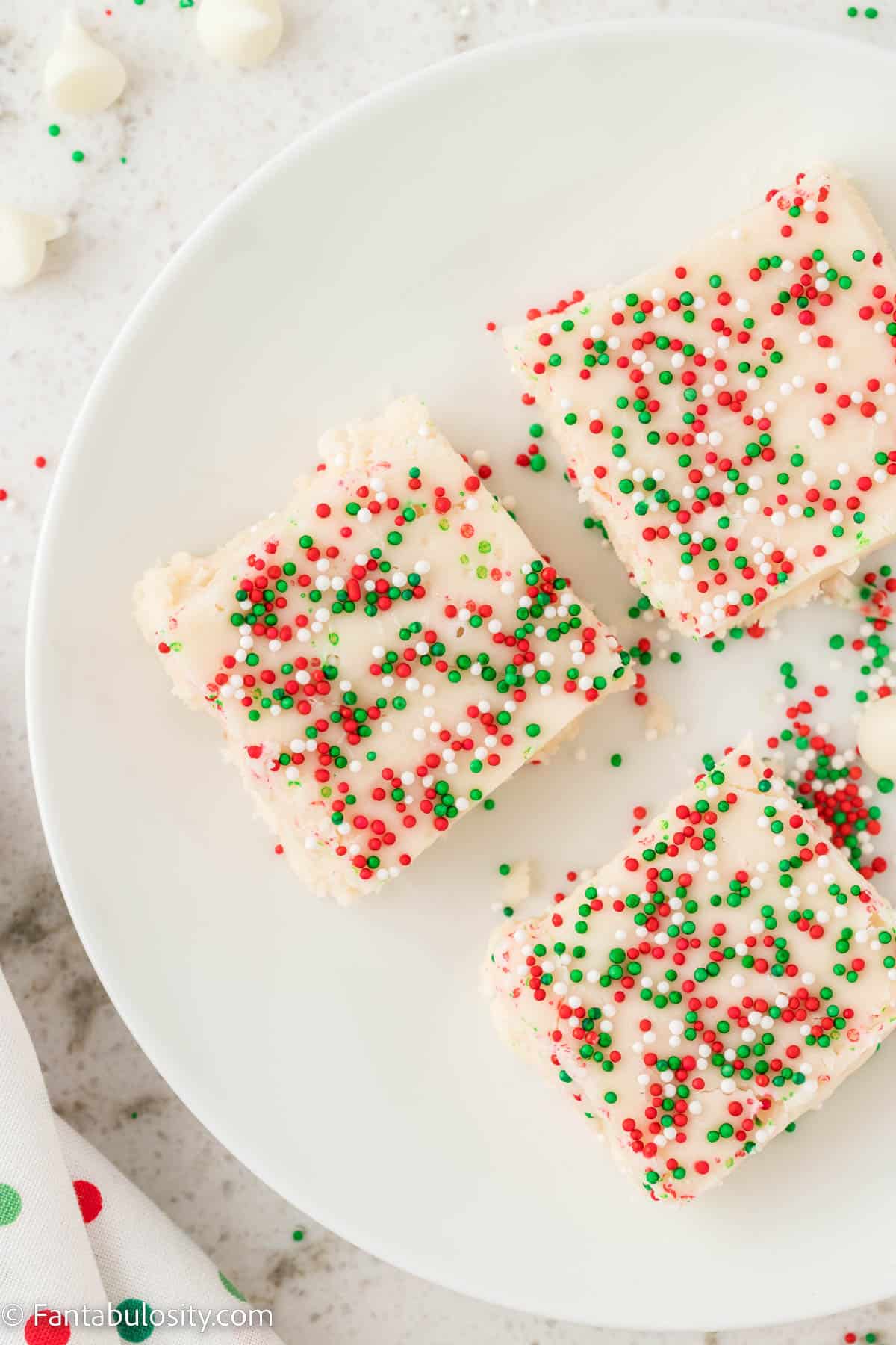 A birdseye photo of a white plate holding three pieces of white fudge topped with red white and green sprinkles