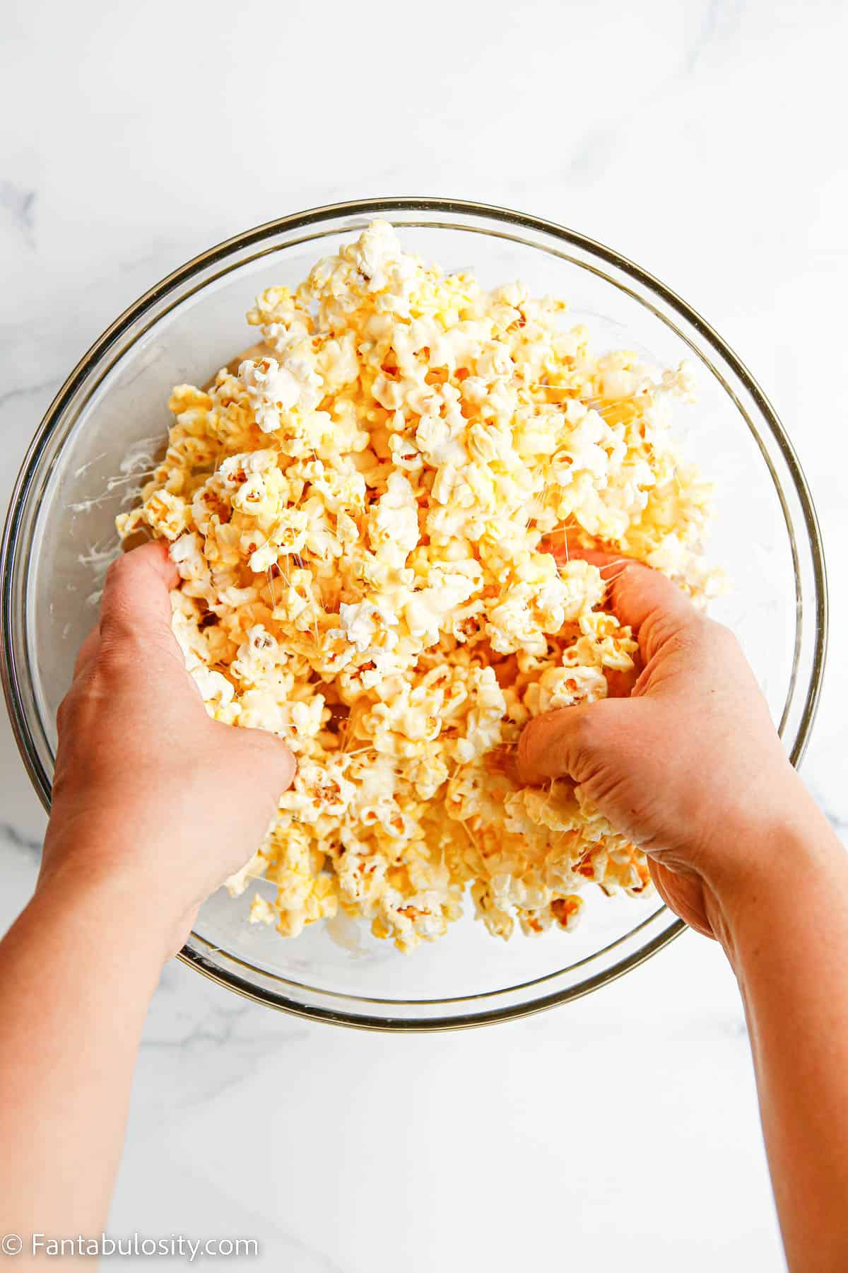 Two hands are mixing together a bowl of popped popcorn and melted marshmallow