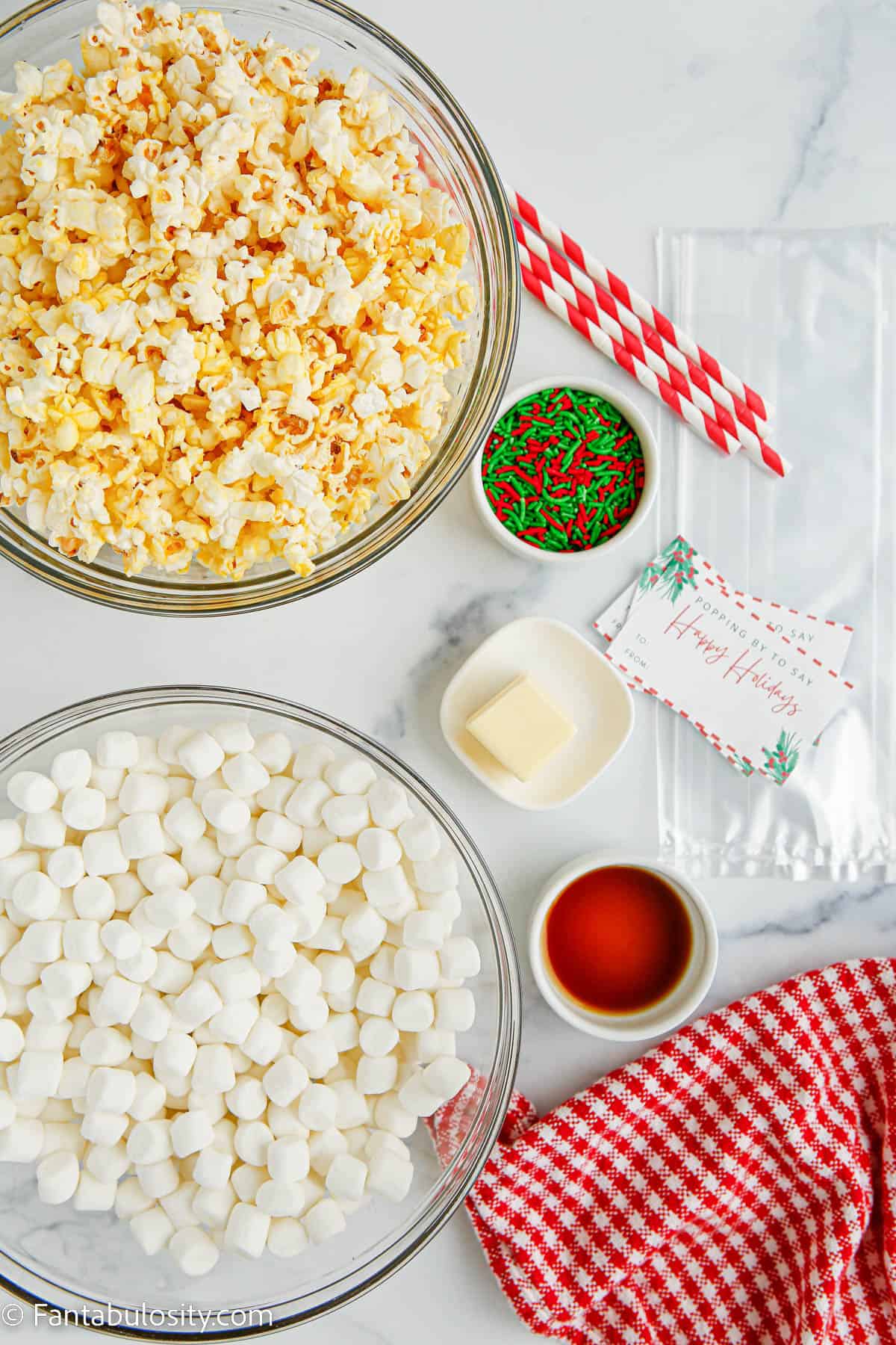 Bowls of popcorn, mini marshmallows, butter, sprinkles and vanilla extract and wrapping supplies are displayed on a white marble background