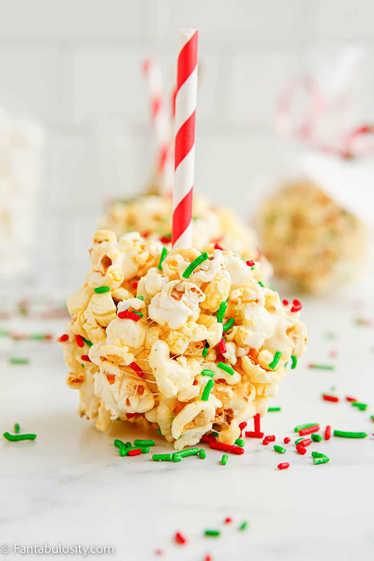 A red and white paper straw has been cut in half and inserted into the center of a sprinkle covered marshmallow popcorn ball