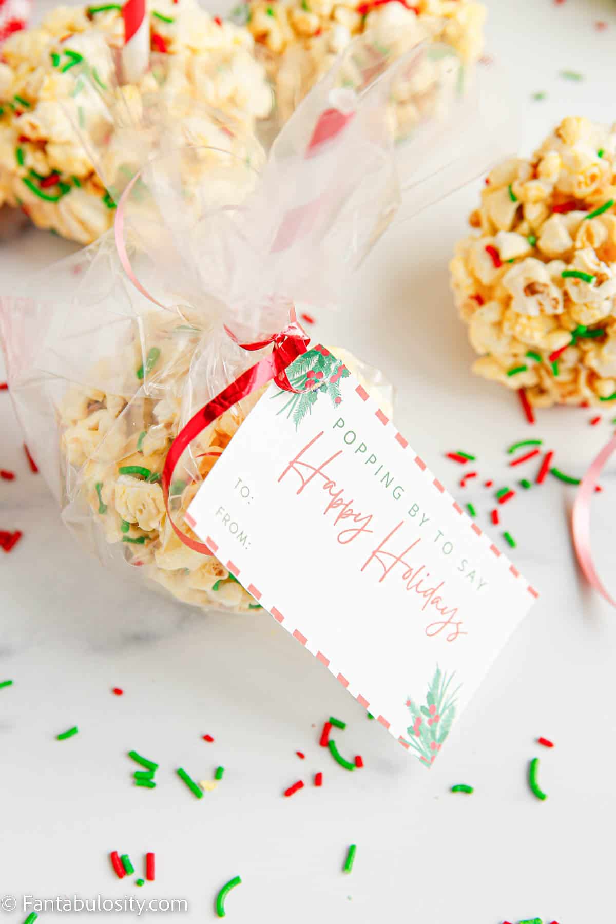 A marshmallow popcorn ball has been placed in a cellophane bag that is tied closed with a red ribbon and a festive holiday gift tag