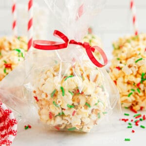 A popcorn ball that has been rolled in red and green sprinkles has been placed in a cellophane bag that is tied closed with red ribbon.