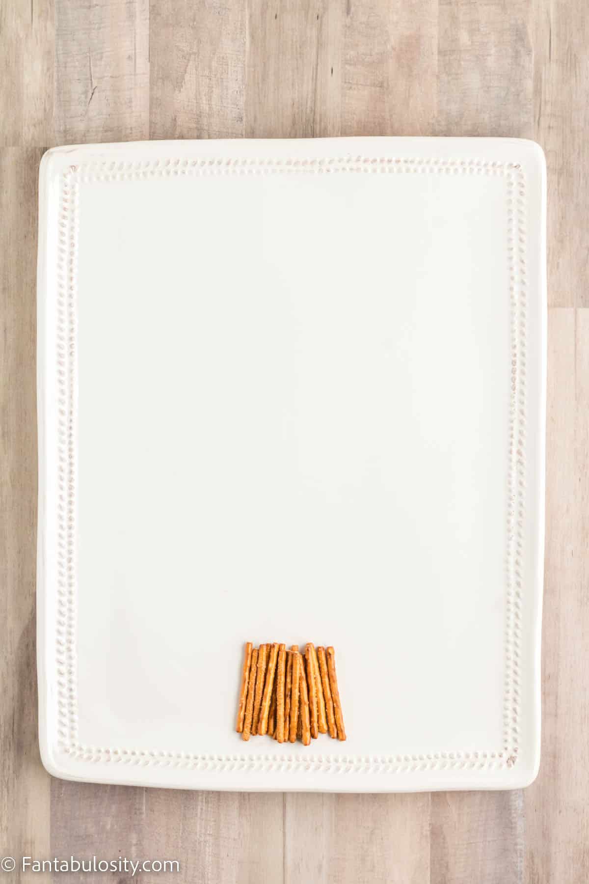 A large rectangular white platter has pretzel sticks together forming the trunk of a Christmas tree charcuterie board