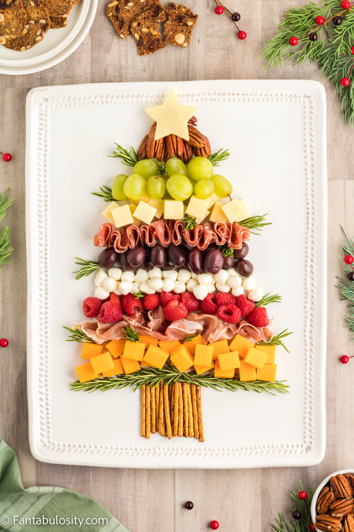 Nuts, fruits, meats and cheeses have been arranged in the shape of a Christmas tree on a white rectangular serving platter