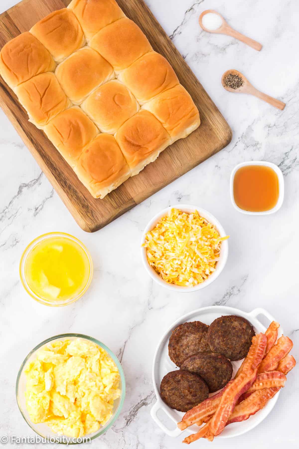Hawaiian rolls, cooked bacon and sausage, scrambled eggs and shredded cheese are displayed on a white marble background