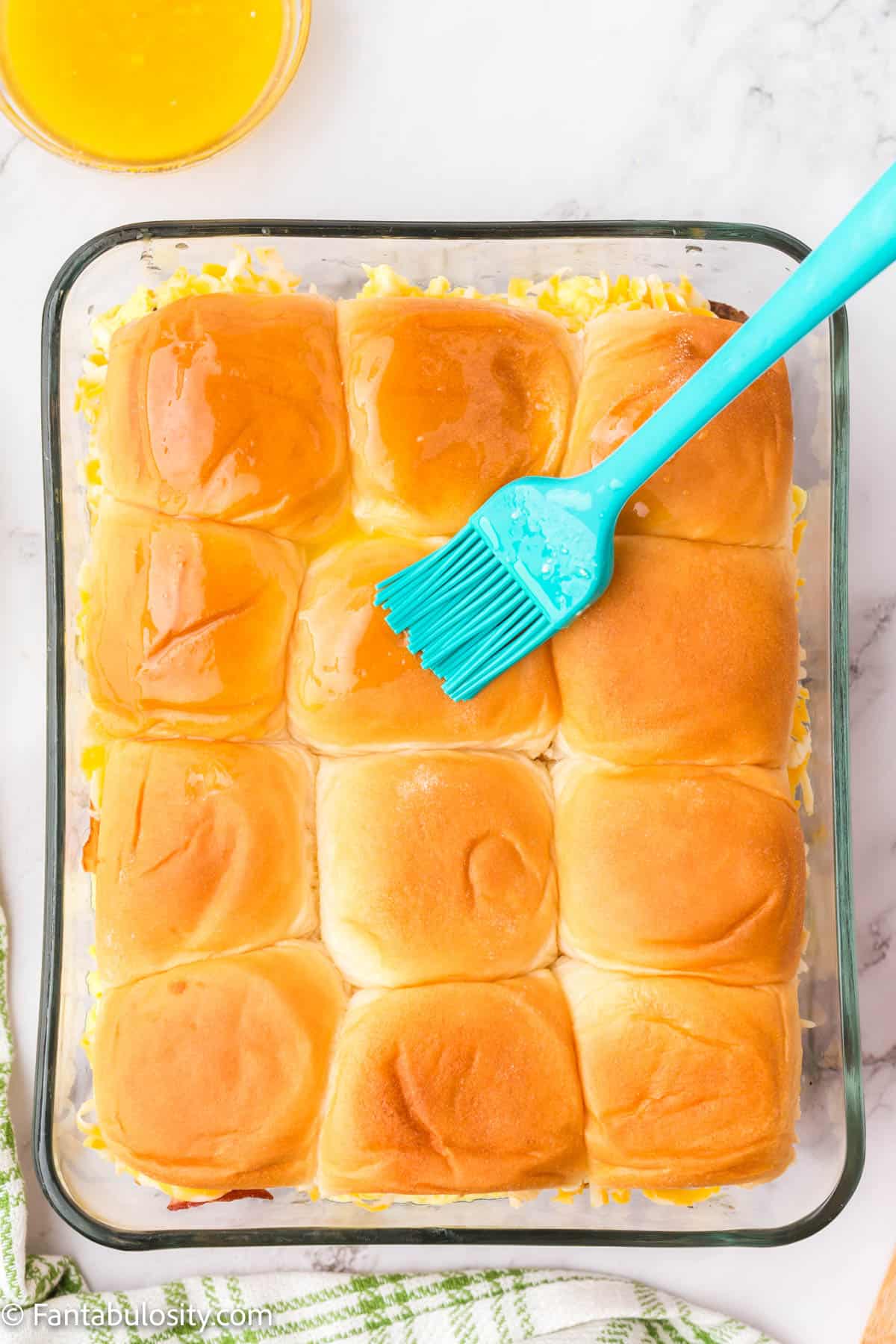 A silicone brush is brushing melted butter over the tops of 12 breakfast sliders in a glass baking dish