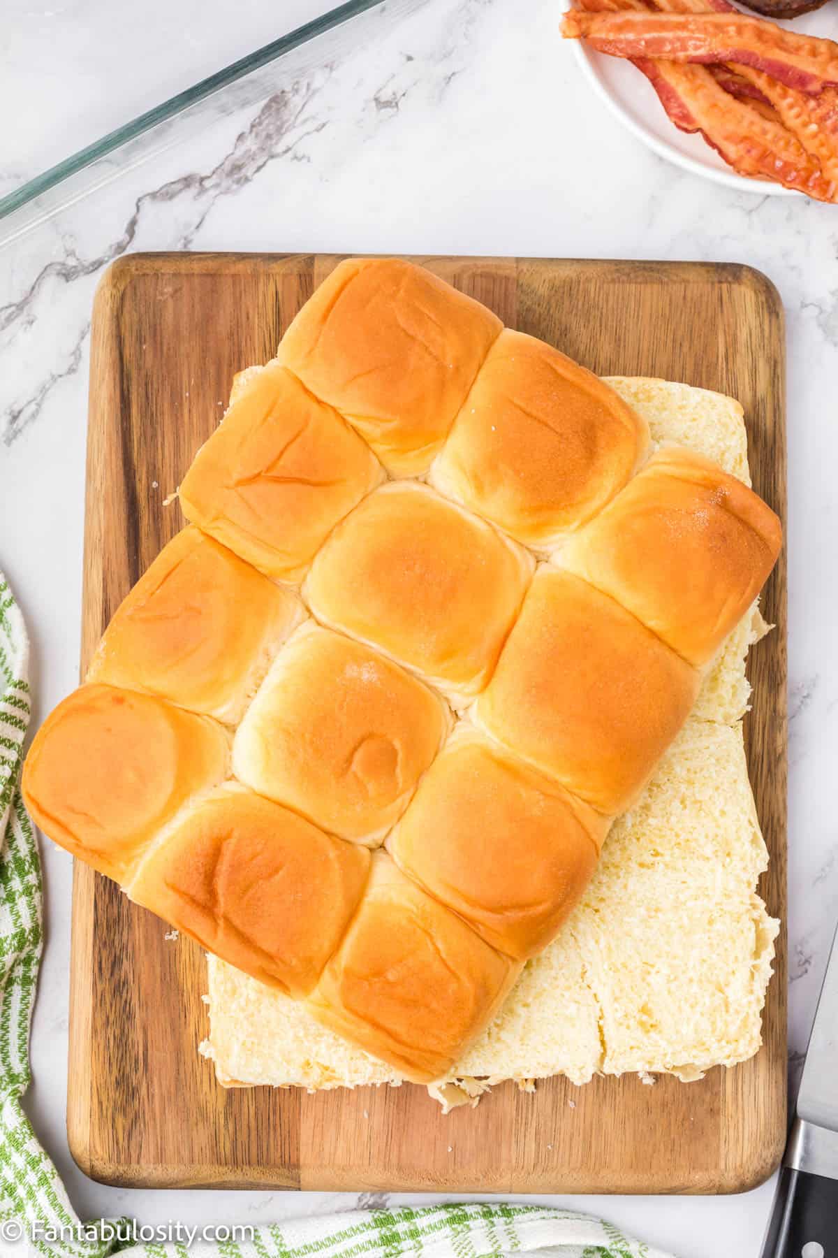 12 Hawaiian rolls have been cut horizontally and are resting on a wooden cutting board