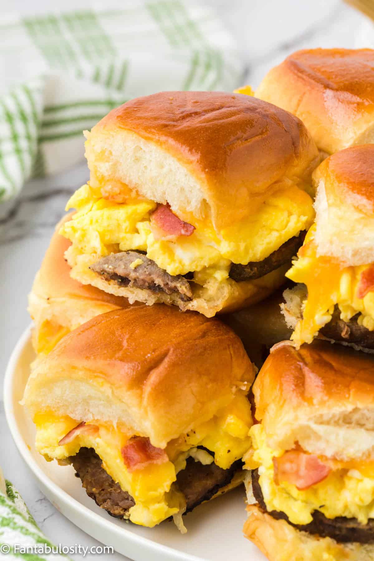 Breakfast slider sandwiches are stacked on a plate making it easy to see sausage, bacon, eggs and cheese on toasted Hawaiian rolls