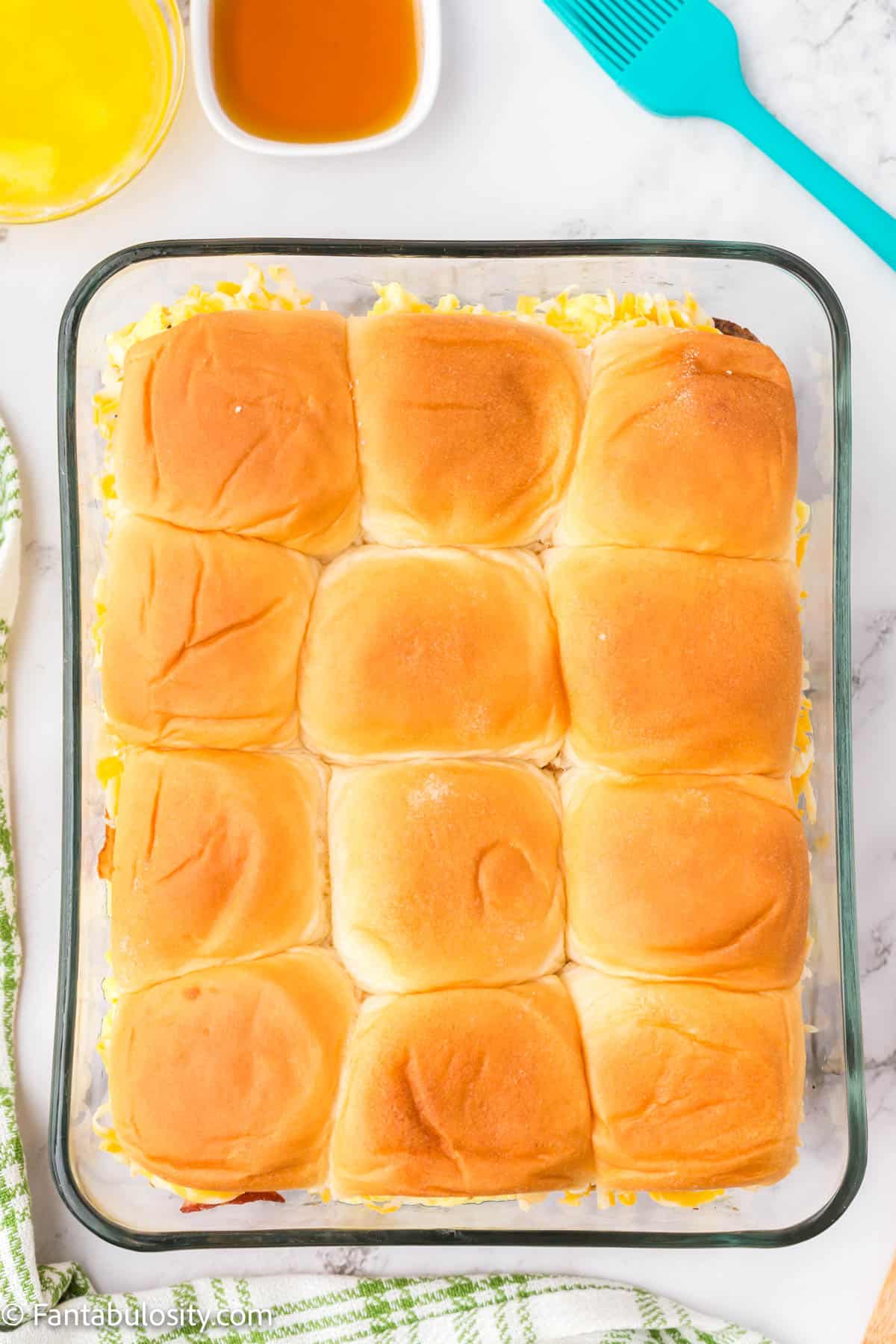 The top buns of 12 breakfast sliders have been placed on top of the eggs, sausage, bacon and cheese in a large glass baking dish