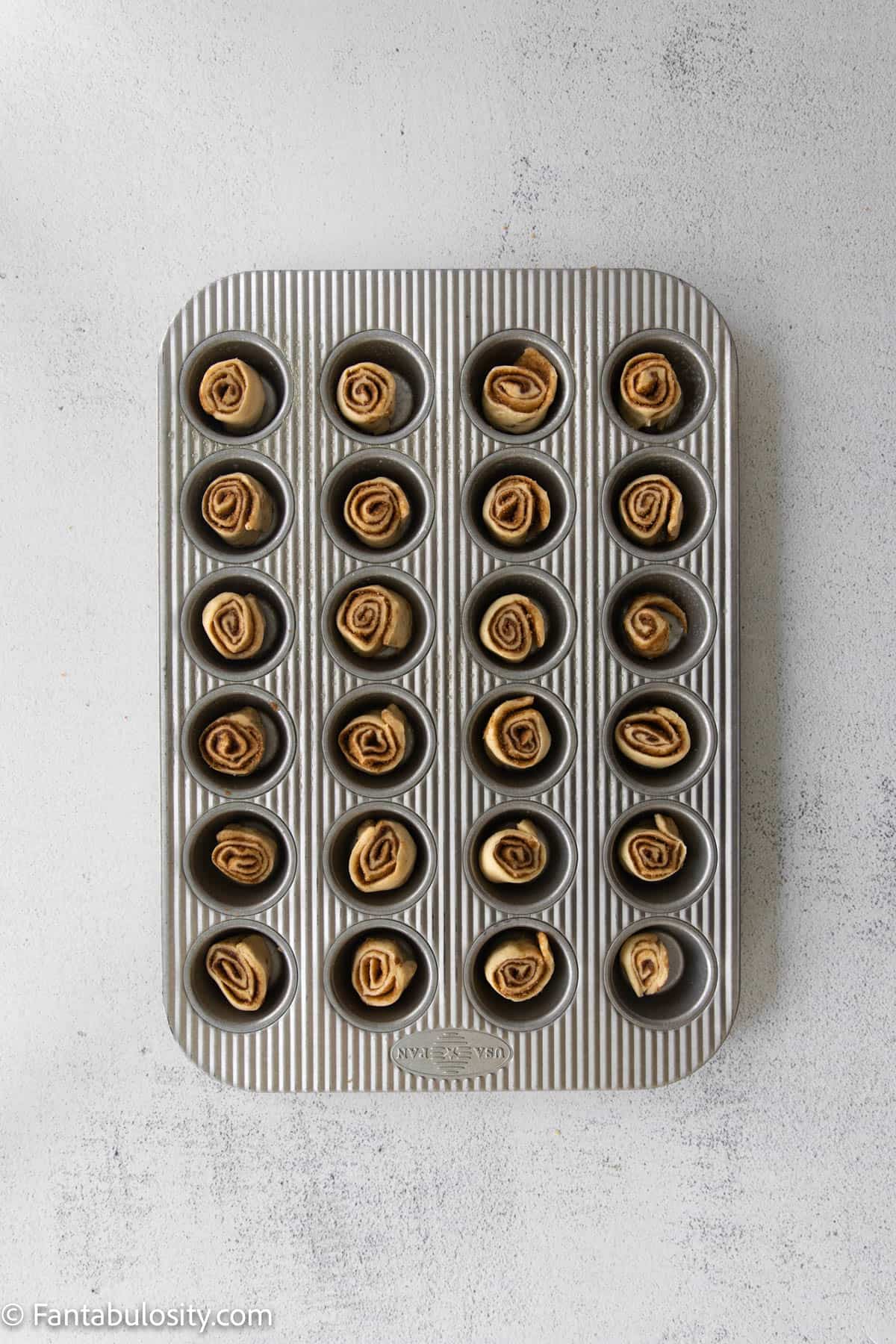 I mini cupcake pan with 24 spaces, each filled with an unbaked mini cinnamon roll.