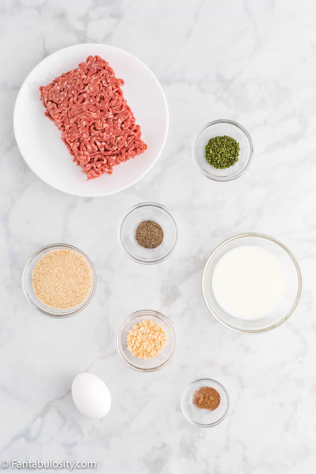Ground beef, an egg, bowls of spices and milk are displayed on a white marble background