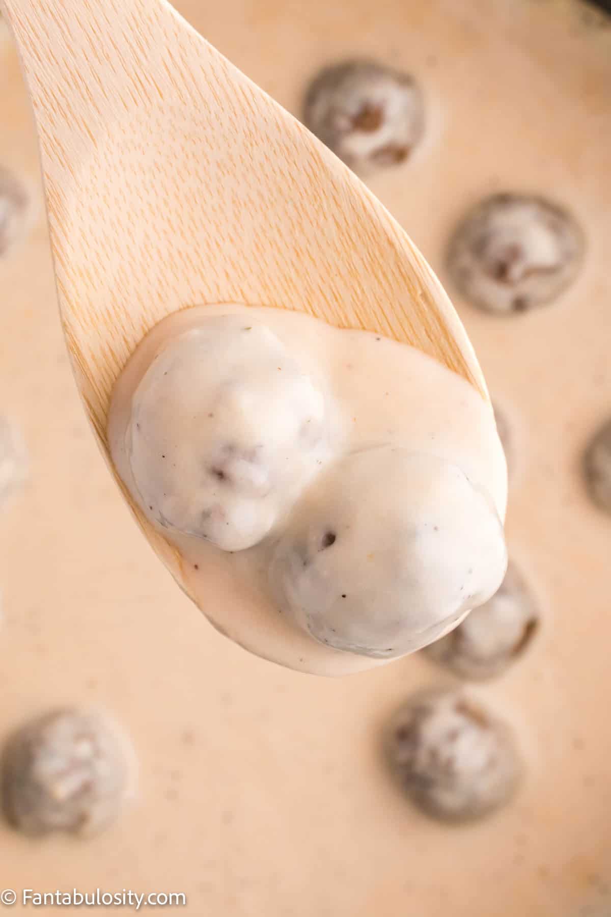 A wooden spoons holds two meatballs and a rich cream sauce above a pan containing the sauce and more meatballs