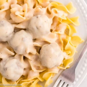 Close up photo of a white plate full of cooked egg noodles topped with meatballs in a creamy sauce