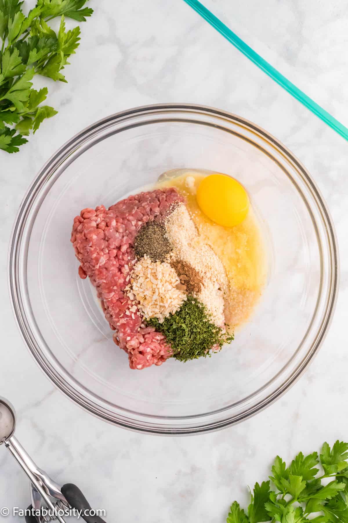 A glass mixing bowl holds ground beef, bread crumbs, an egg and spices to mix to make meatballs