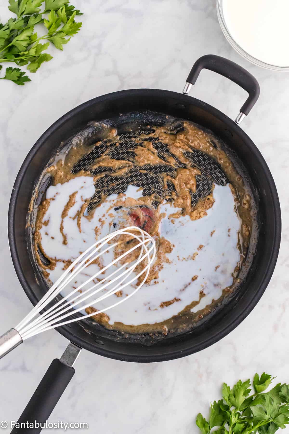 A whisk is poised above a large black skillet that holds the beginning of a cream sauce