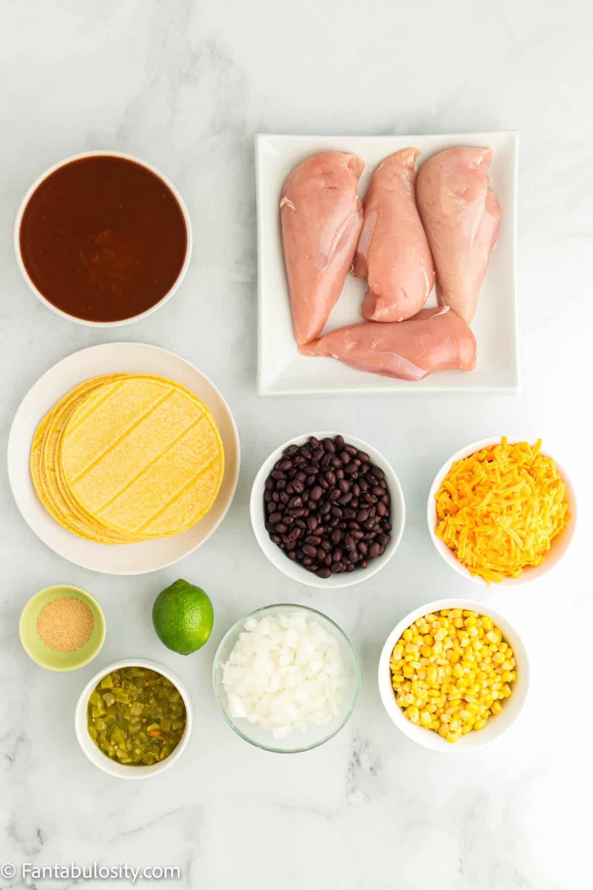 Corn tortillas, chicken breasts, enchilada sauce, corn, black beans, green chiles, onions, cheese and a lime are displayed on a white marble background