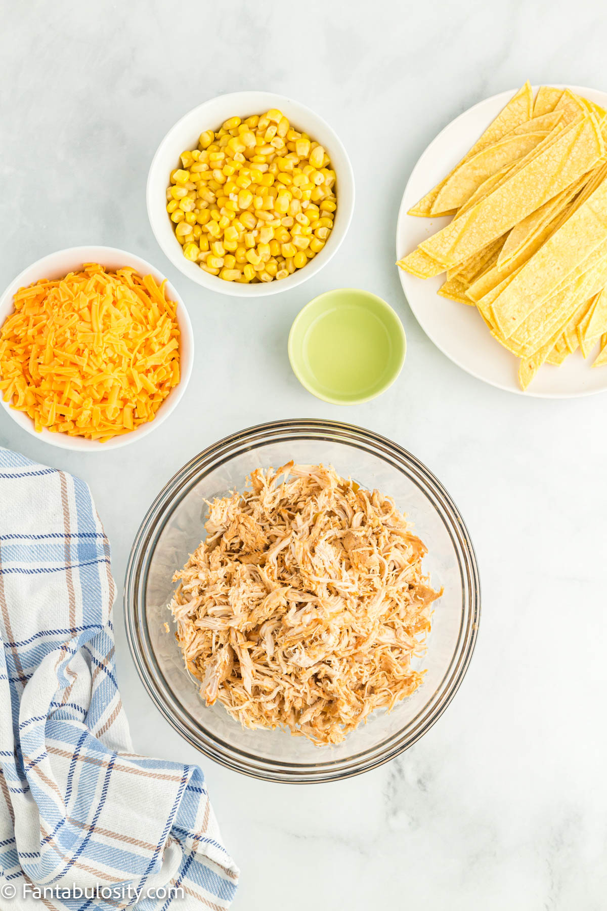 Bowls hold shredded chicken, corn, shredded cheese and lime juice and a plate of corn tortillas are also displayed on a white marble background