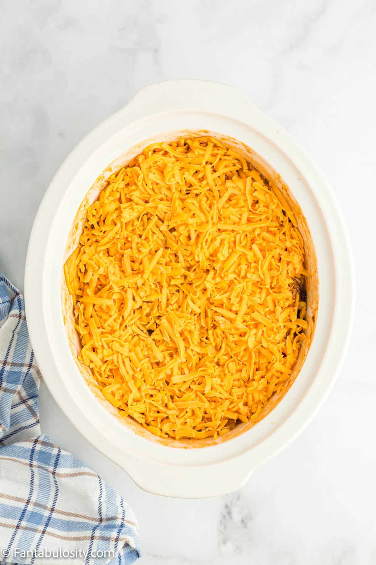 A slow cooker insert holds a mixture of chicken enchilada filling that has been covered with a layer of shredded cheddar cheese