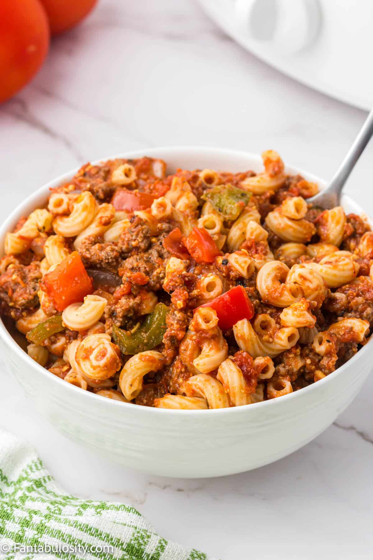 A spoon is tucked into a white bowl filled to the brim with goulash packed with vegetables, beef and noodles