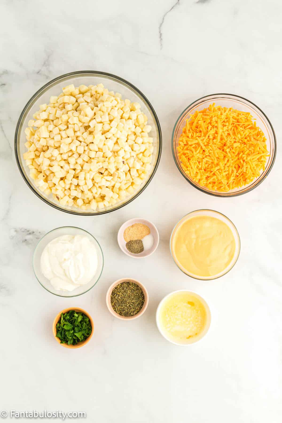 Diced hashbrown potatoes, shredded cheddar cheese and several other bowls of ingredients are displayed on a white marble background
