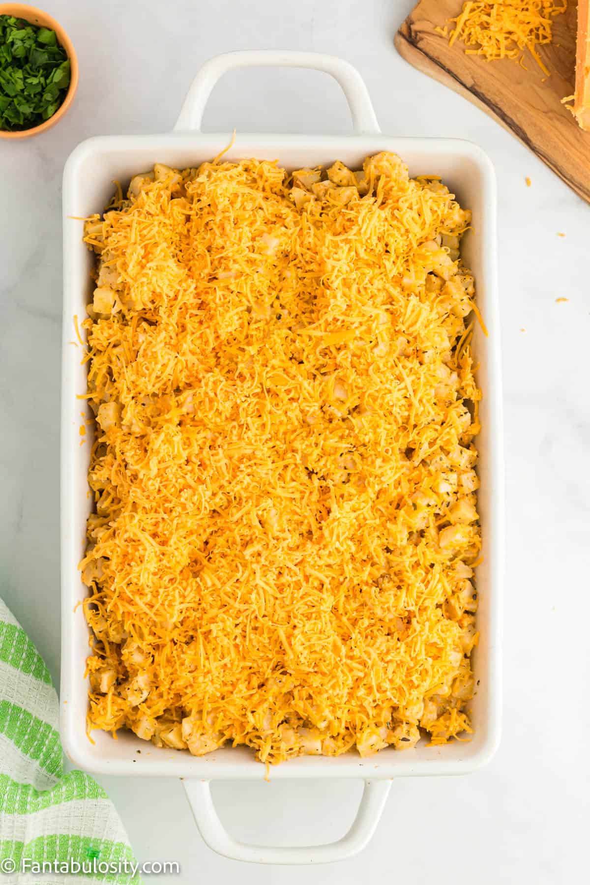 A white 9x13 baking dish holds seasoned diced hashbrown potatoes that have been topped with shredded cheddar cheese