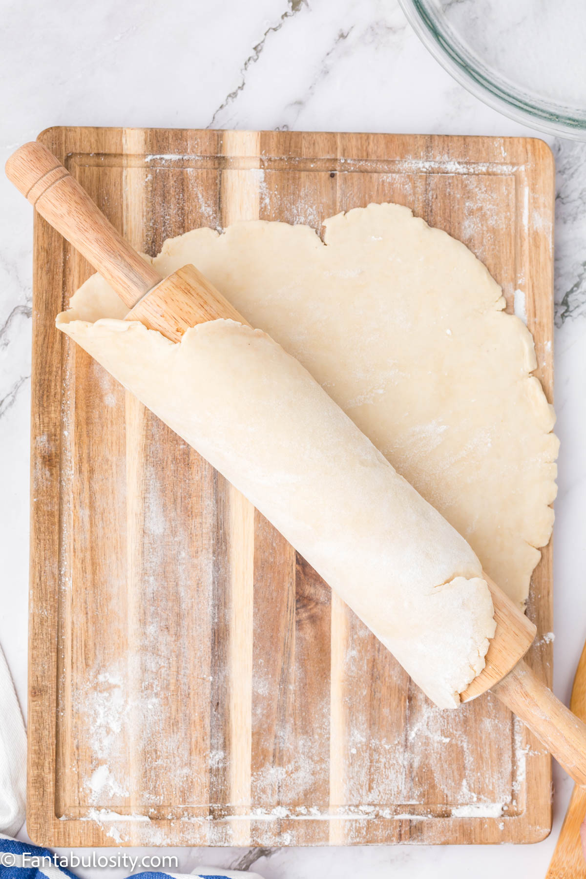 Pie dough being rolled from a wood cutting board onto a wooden rolling pin.