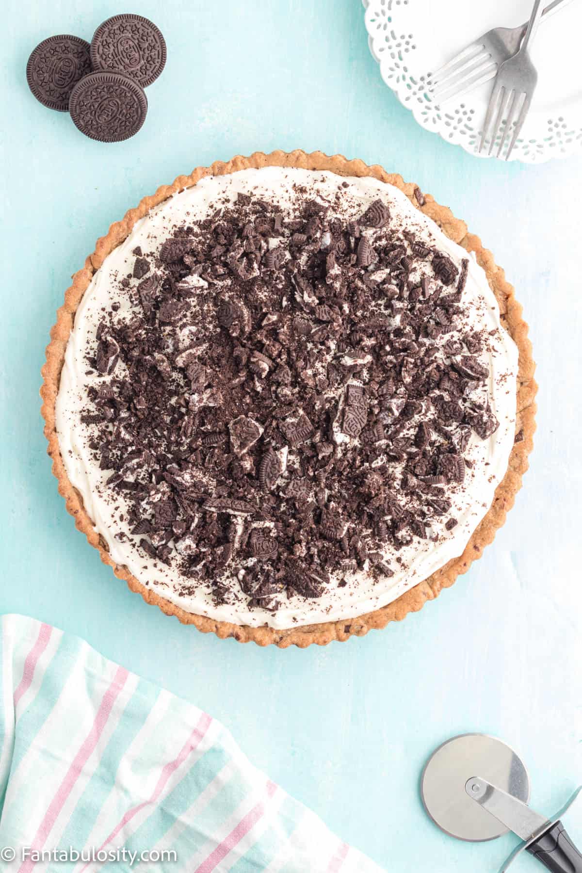 A round Oreo dessert pizza is in center of the photo while plates and forks and a pizza wheel wait nearby to serve it with