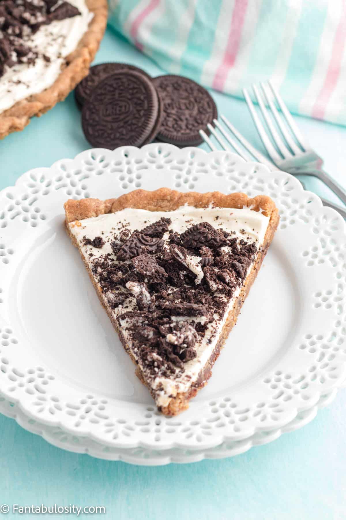 A slice of Oreo pizza rests on a pretty white plate and the rest of the pizza, a few Oreo cookies and forks are visible in the background