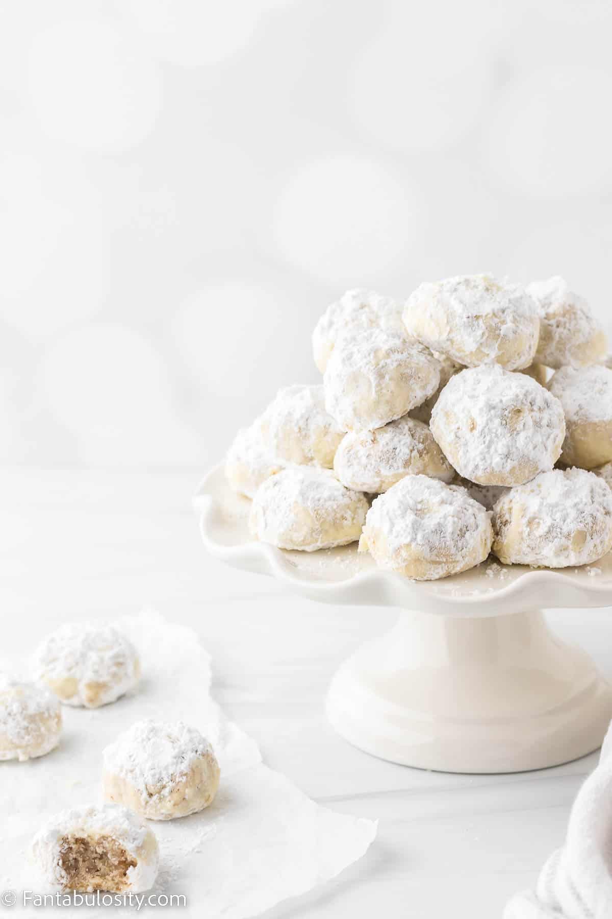 Pecan snowball cookies have been piled on a white cake stand and several additional cookies, one with a bite gone, are sitting on a piece of white parchment paper